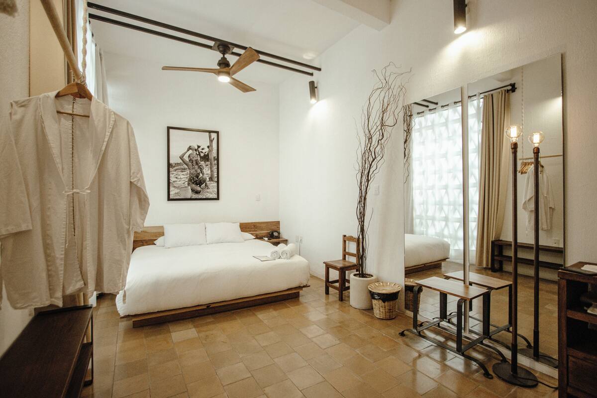Un dormitorio con cama, cabecera de madera, ventilador de techo, mesita de noche, silla, planta en maceta, espejo y bata colgada.