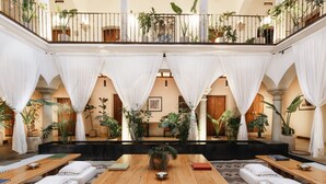 Un amplio atrio con una mesa de madera en el centro, rodeada de asientos acolchados, plantas en macetas y un balcón con barandillas.