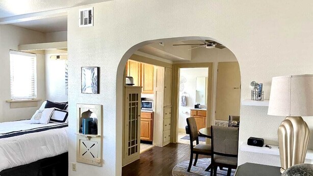 A bedroom with a bed, a desk, and a chair. There is a kitchen area visible through an archway. - Evie's apartment (Oklahoma City)