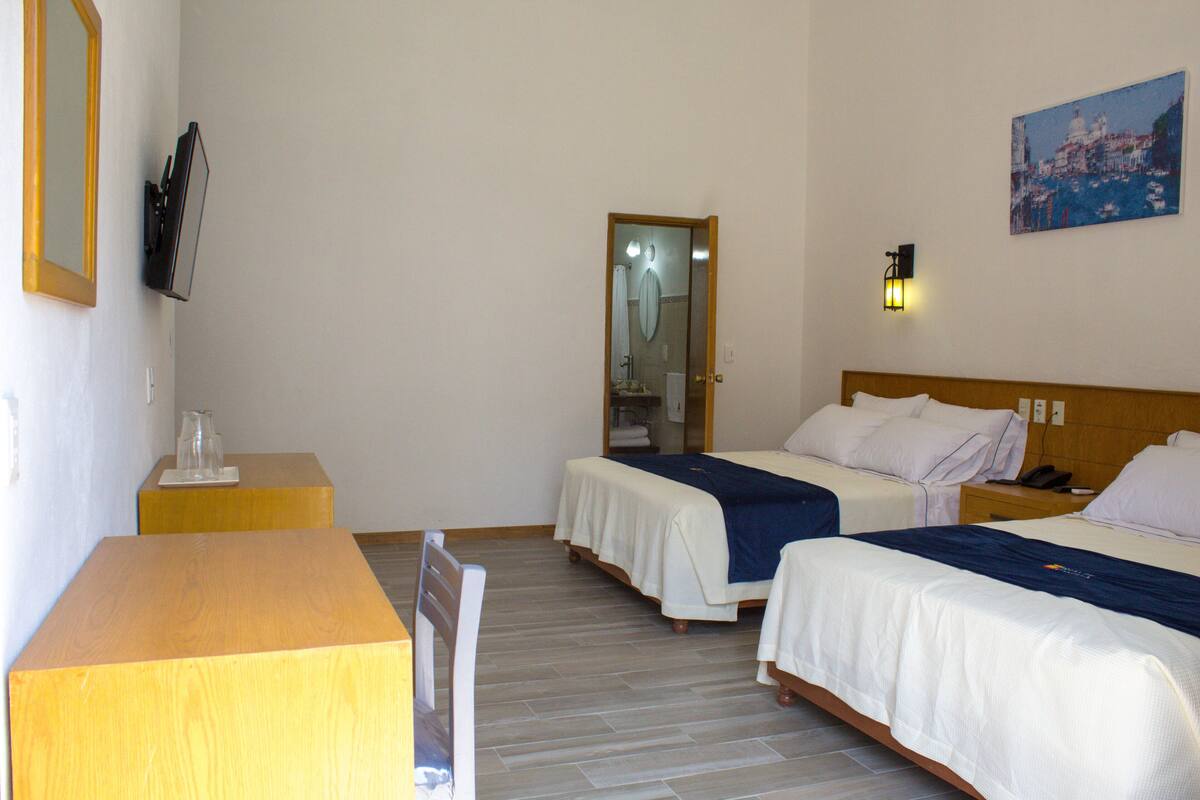 Habitación de hotel con dos camas, un escritorio de madera, un televisor y un cuadro en la pared.
