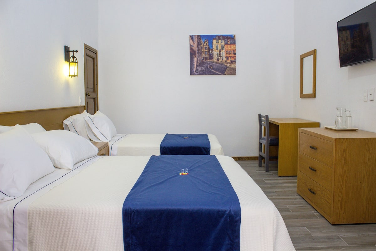 Habitación de hotel con dos camas, un aparador de madera, un escritorio, una silla, un televisor y un cuadro en la pared.