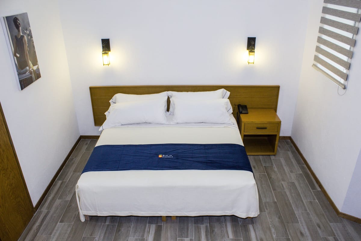 Una cama bien hecha con sábanas blancas, un cabecero de madera y dos mesitas de noche con lámparas fijadas a la pared.
