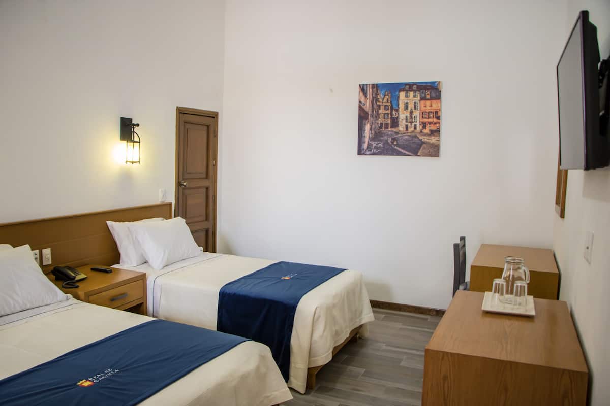 Habitación de hotel con dos camas, una mesita de noche de madera con un teléfono, una mesita de noche con una jarra de vidrio y un televisor montado en la pared.