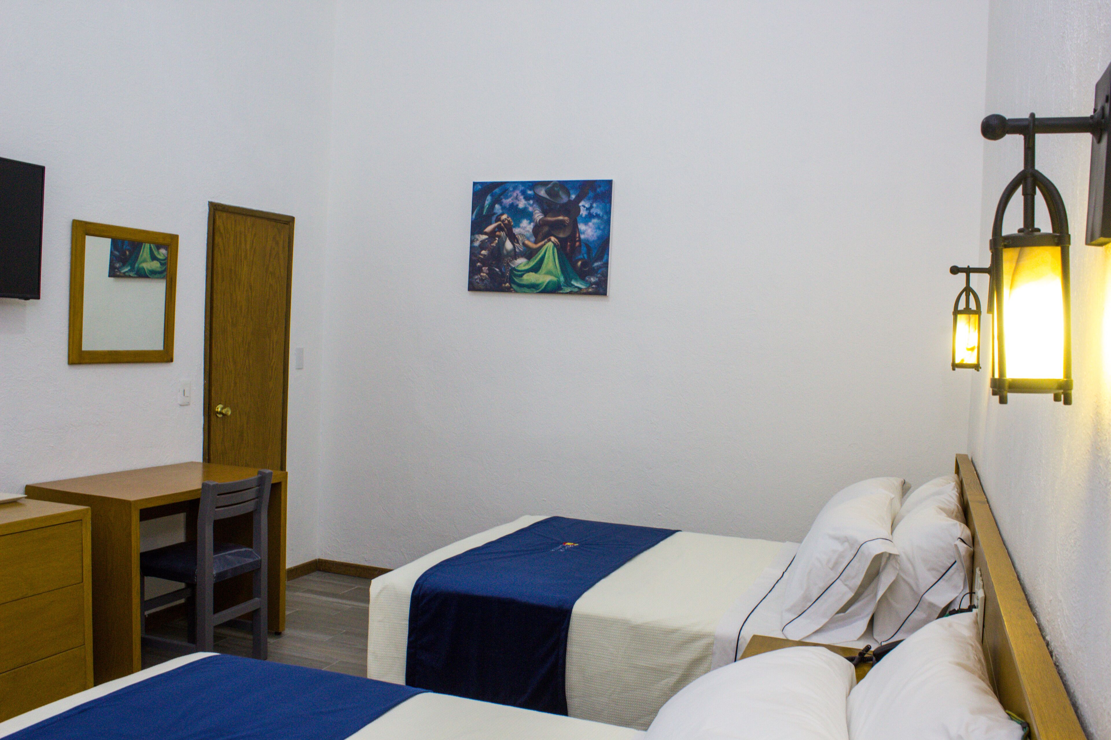 A hotel room with two beds, a desk, a chair, a wardrobe, and artwork on the wall.