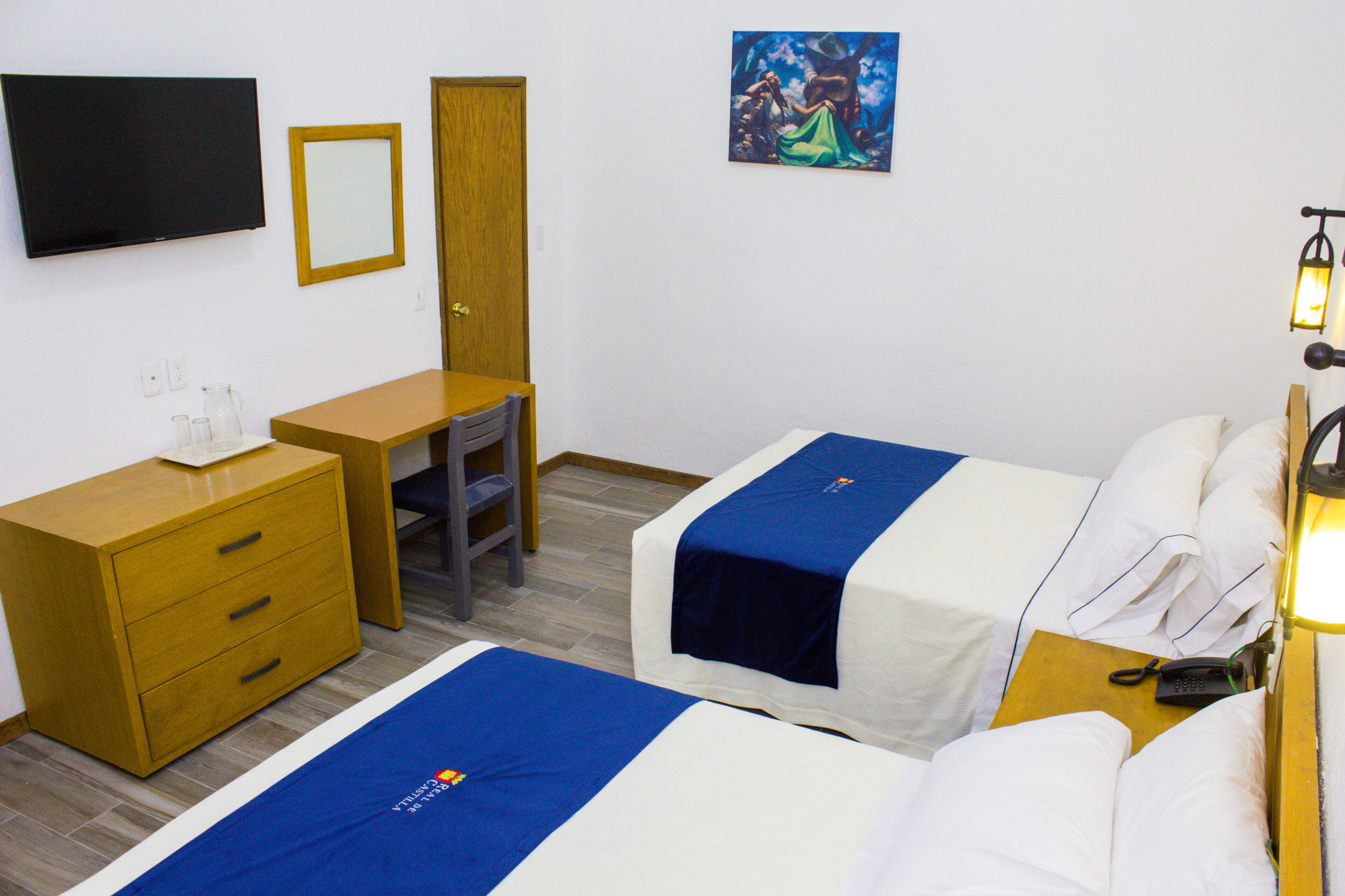 Hotellihuone, jossa on kaksi sänkyä, työpöytä, tuoli, televisio, taulu ja yöpöytälamppu.