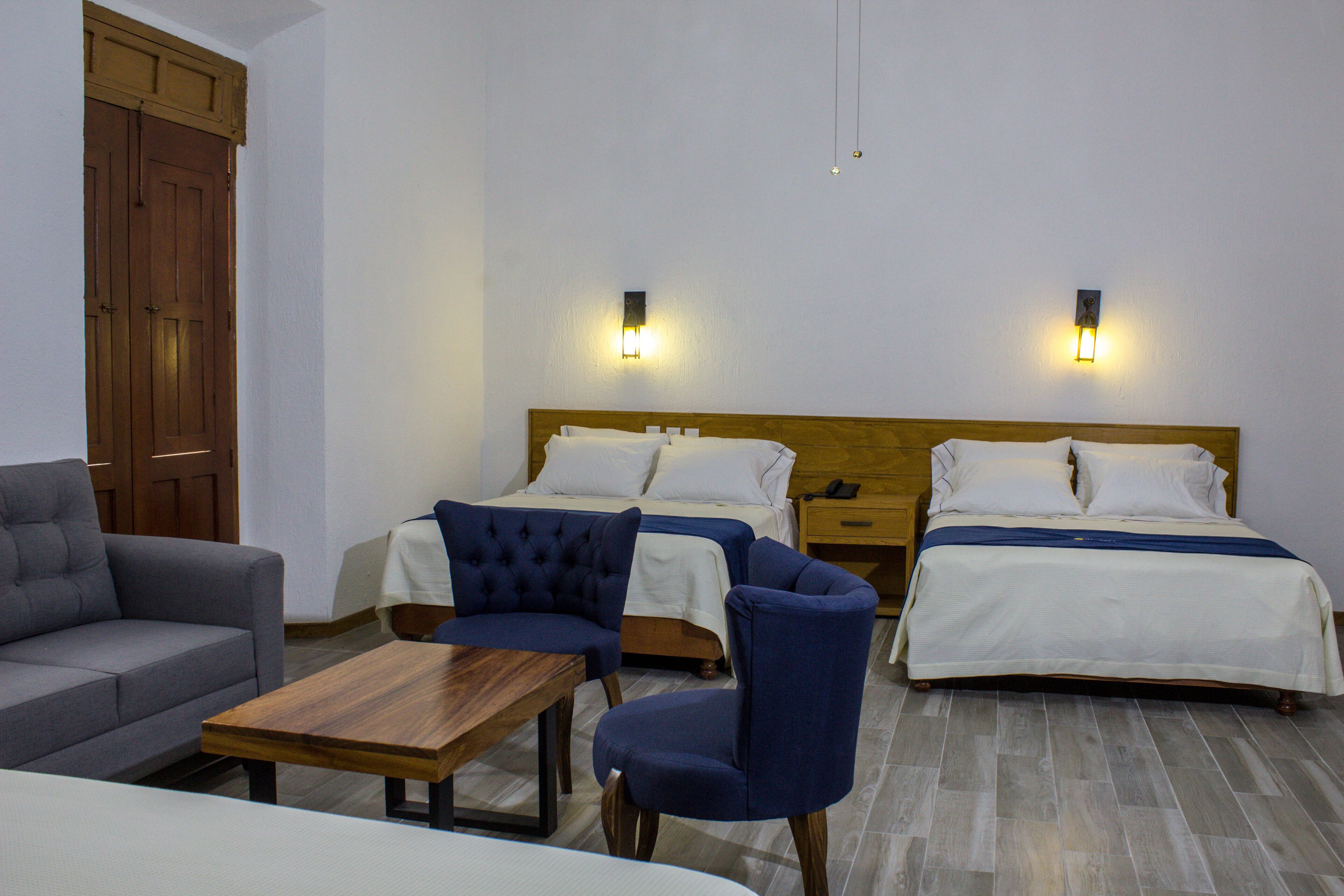 A hotel room with two beds, a sofa, a table, and two wall-mounted lights.