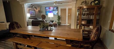 Uma área de refeições com mesa e bancos de madeira, ventoinha no teto, televisão e armário com portas de vidro.