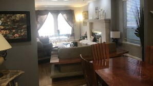 Uma sala de estar com sofá, mesa de jantar com cadeiras, lareira e uma janela grande com cortinas.