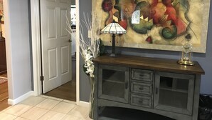 Um corredor com porta dando para outro quarto, uma pintura abstrata pendurada na parede, um abajur, um vaso com flores e um relógio em um armário.