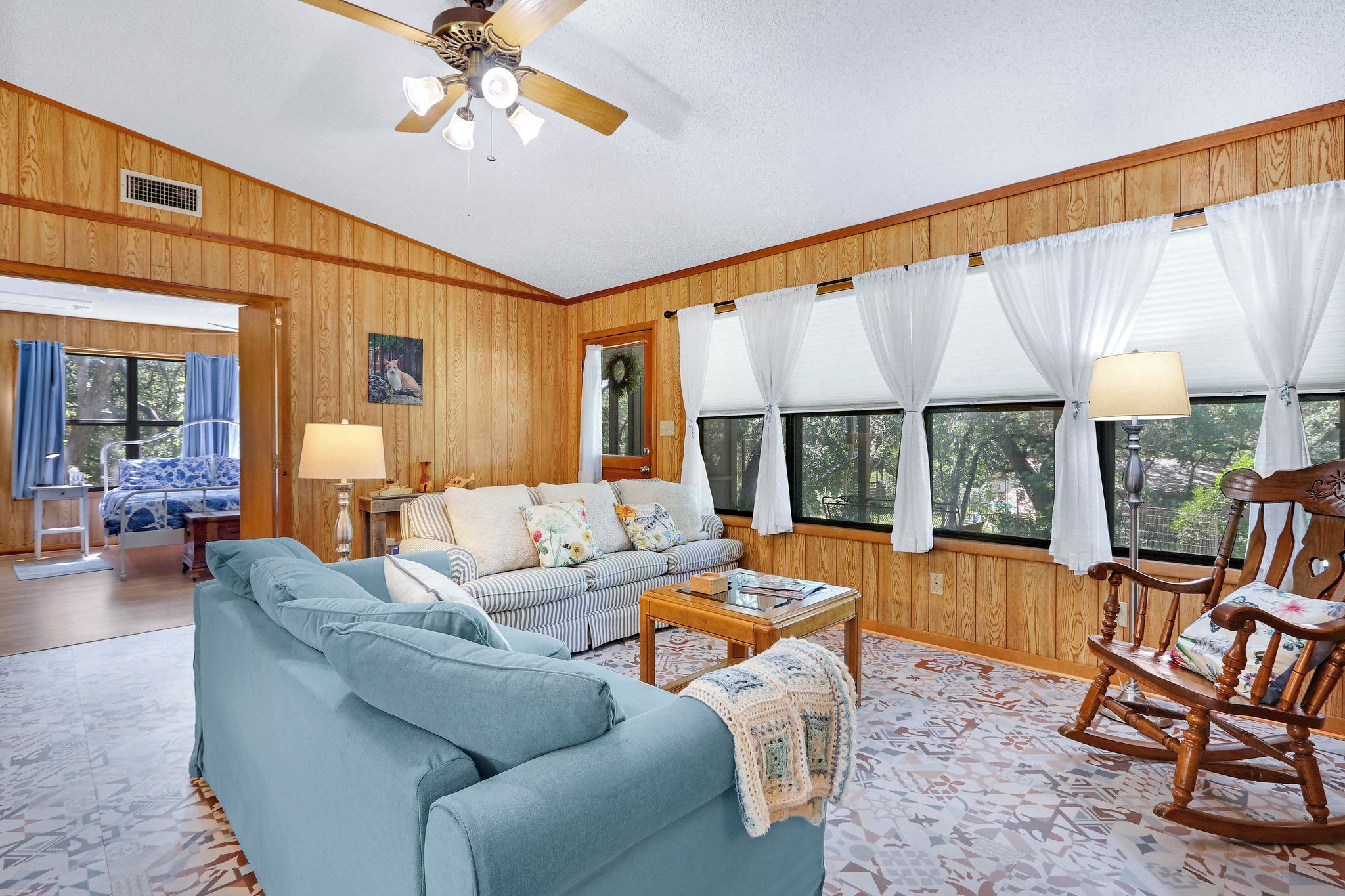 A room with a wooden paneled wall, a ceiling fan, a sofa, a rocking chair, and a window with curtains.