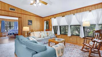 A room with a wooden paneled wall, a ceiling fan, a sofa, a rocking chair, and a window with curtains.