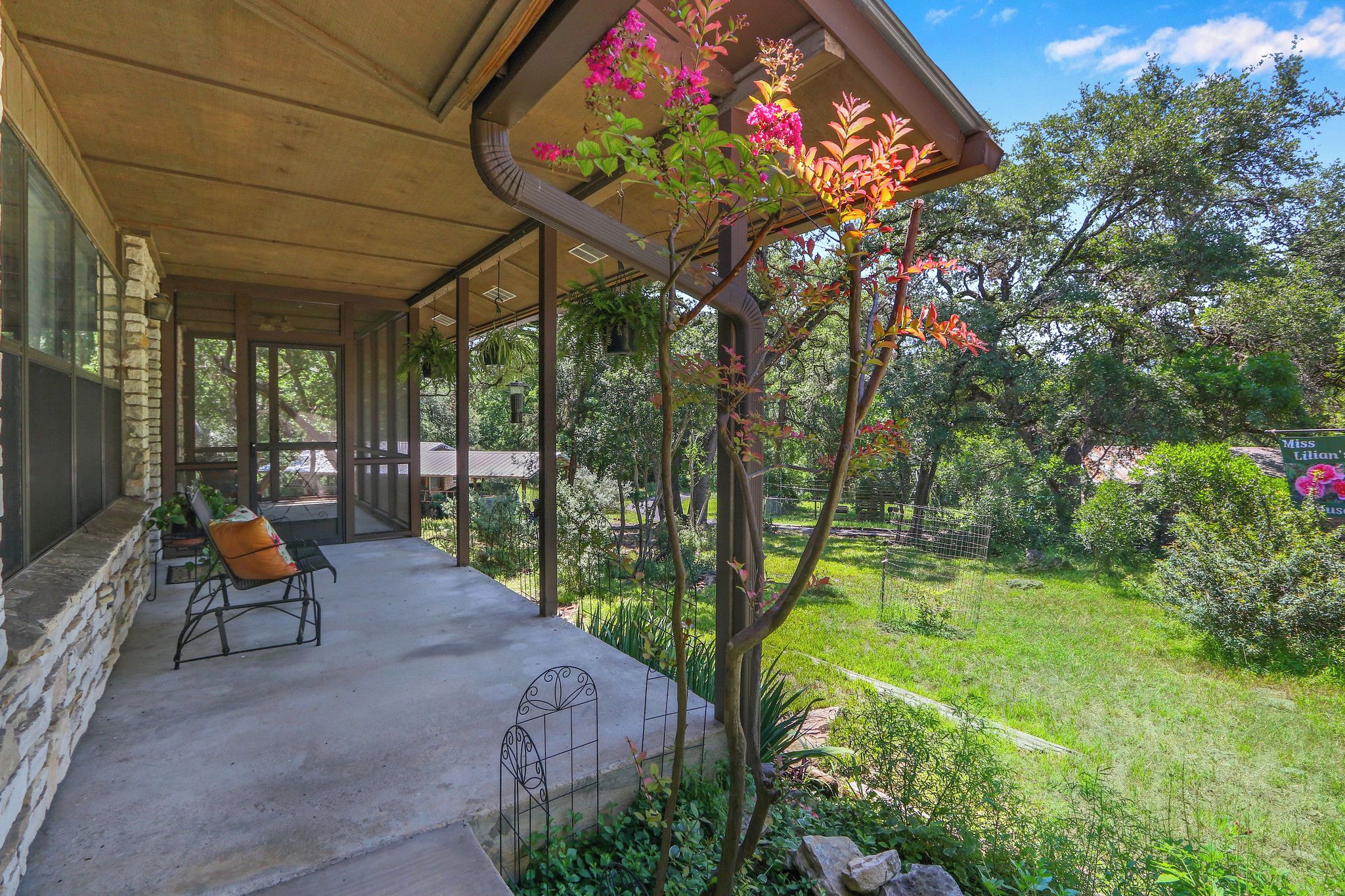 A covered outdoor seating area with a stone wall, a metal chair, and a garden with various plants and a tree with pink flowers.