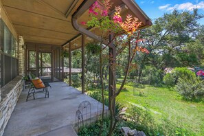 A covered outdoor seating area with a stone wall, a metal chair, and a garden with various plants and a tree with pink flowers.