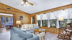 Une pièce avec un ventilateur de plafond, un canapé, un fauteuil à bascule et une fenêtre avec des rideaux.