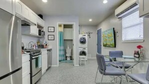 A compact kitchen with a refrigerator, stove, and microwave. There is a dining area with a glass table and chairs. A washing machine is visible in the background. - Garage Studio near OUSHC Hospital (Oklahoma City)