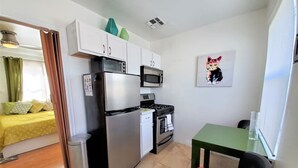A compact kitchen with a refrigerator, microwave, stove, and sink. There is a dining table with a chair, and a bed with a yellow bedspread in the background. - Ar`s Artsy Downtown Bungalow 5 (Phoenix)