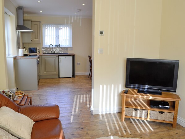 A living room with a brown leather sofa, a television on a wooden stand, and a kitchen in the background. - 3 bedroom accommodation in Talgarth (Talgarth)