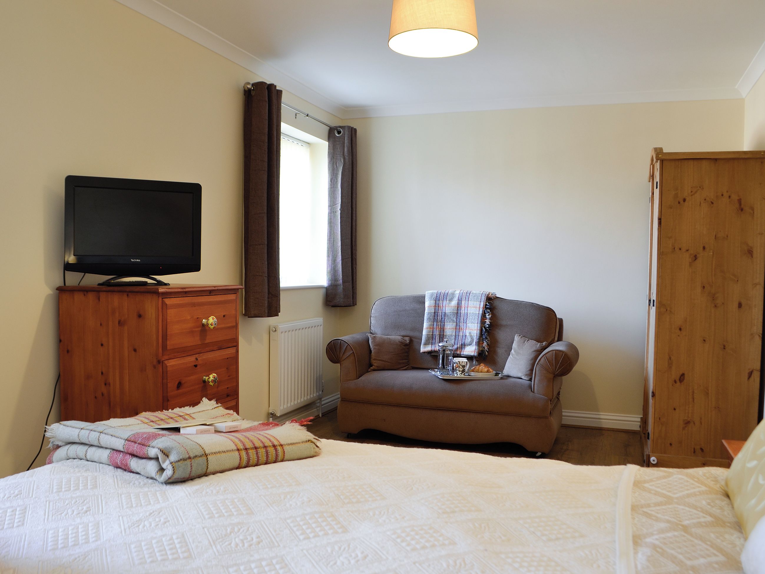 Une chambre à coucher avec un lit, un téléviseur posé sur une commode en bois, un fauteuil marron avec une couverture, et une armoire en bois.