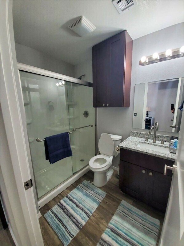 A modern bathroom with a glass shower enclosure, a toilet, a sink with a vanity, and a mirror with lights above. - Private Bedroom Suite - Communal House near Integris Private (Oklahoma City)