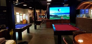 Una sala de billar con varias mesas de billar, una zona de bar y una gran pantalla que muestra un paisaje pintoresco.