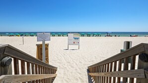 A beach entrance with wooden railings, a signboard, and beach umbrellas in the background.