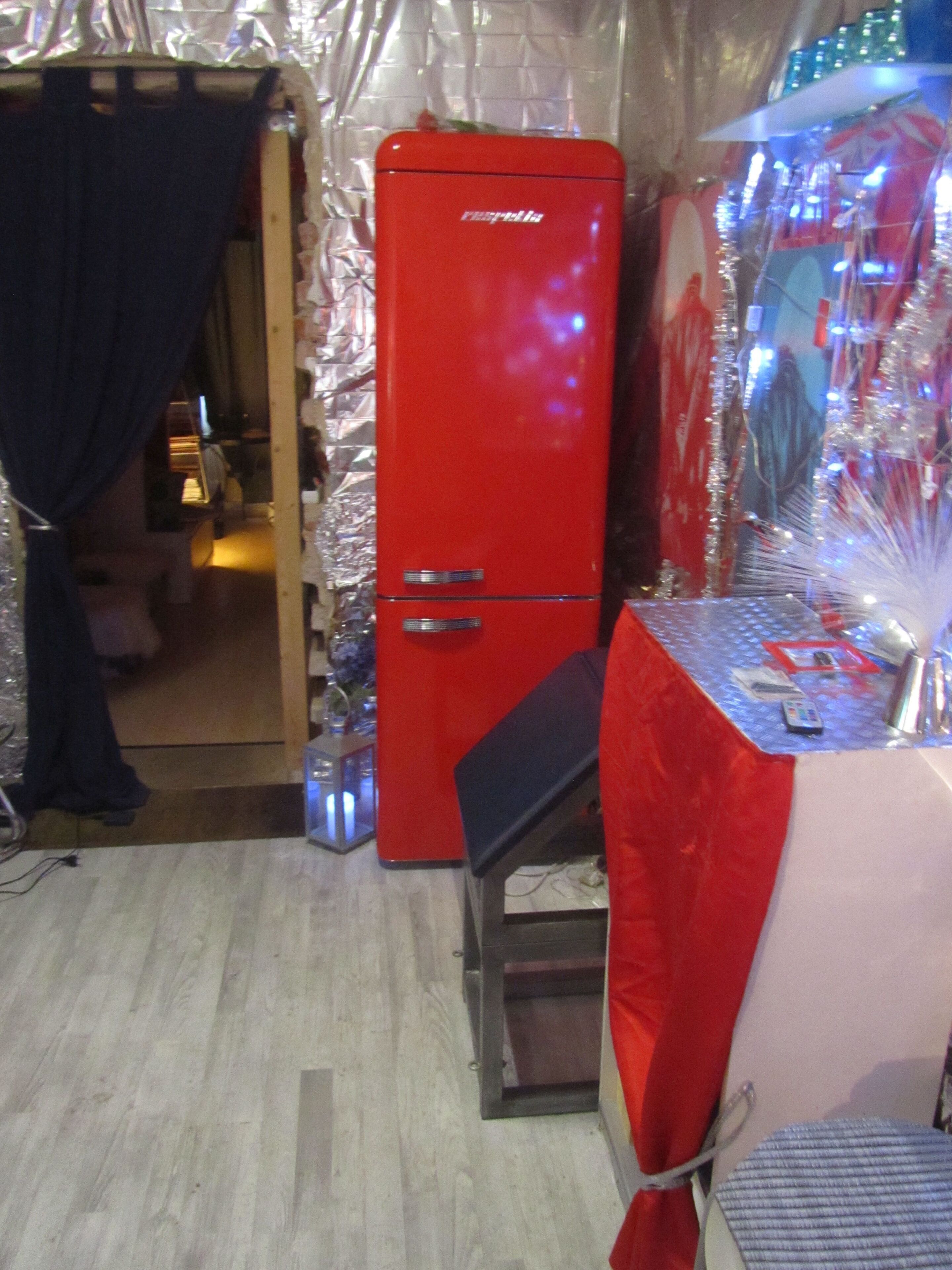 Ein kleines Zimmer mit einem roten Kühlschrank, einem Tisch mit einem Stuhl und einer Tür zu einem weiteren Raum.
