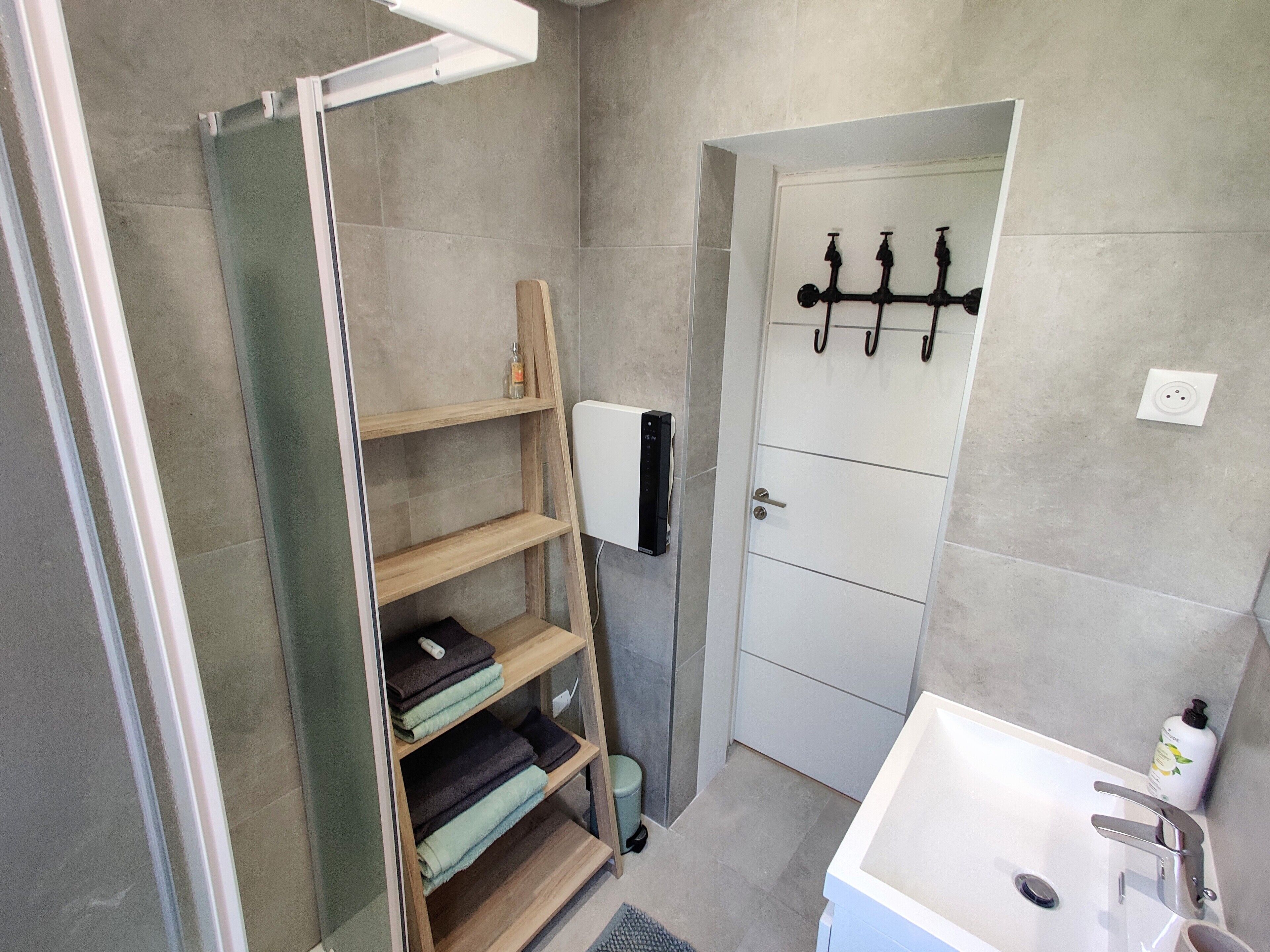 Une salle de bain avec un lavabo blanc, un porte-serviettes, une étagère en bois où sont pliés des serviettes, et une porte dotée d’un distributeur de serviettes.