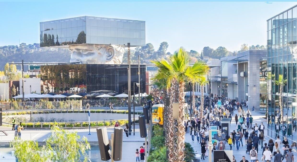 Une place urbaine moderne, avec une fontaine centrale, entourée de palmiers et de passants.