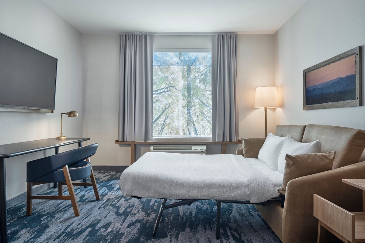A hotel room with a bed, a desk, a chair, a TV, a window with curtains, a lamp, and a painting on the wall.