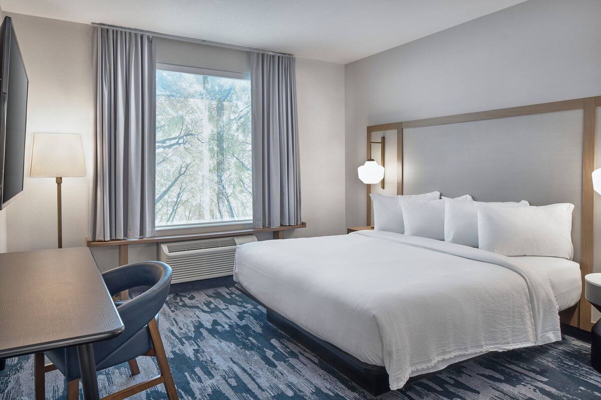 Ein Hotelzimmer mit einem großen Bett, einem Schreibtisch, einem Stuhl und einem Fenster mit Vorhängen.