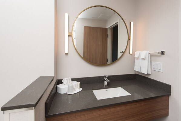 A bathroom with a dark countertop, a white sink, a round mirror, and two wall-mounted towel racks. - Fairfield Inn & Suites by Marriott Penticton (Penticton)