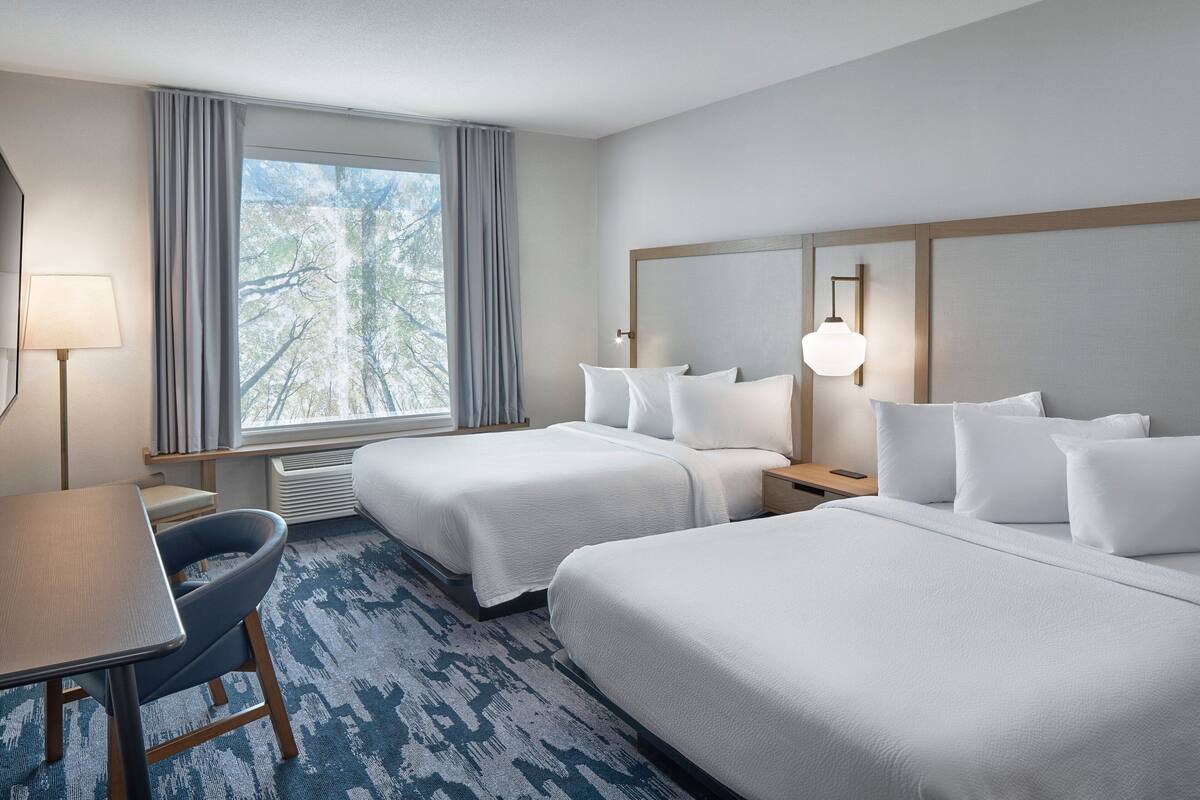 Ein Hotelzimmer mit zwei Betten, einem Schreibtisch, einem Stuhl, einer Lampe und Blick auf Bäume durch das Fenster.
