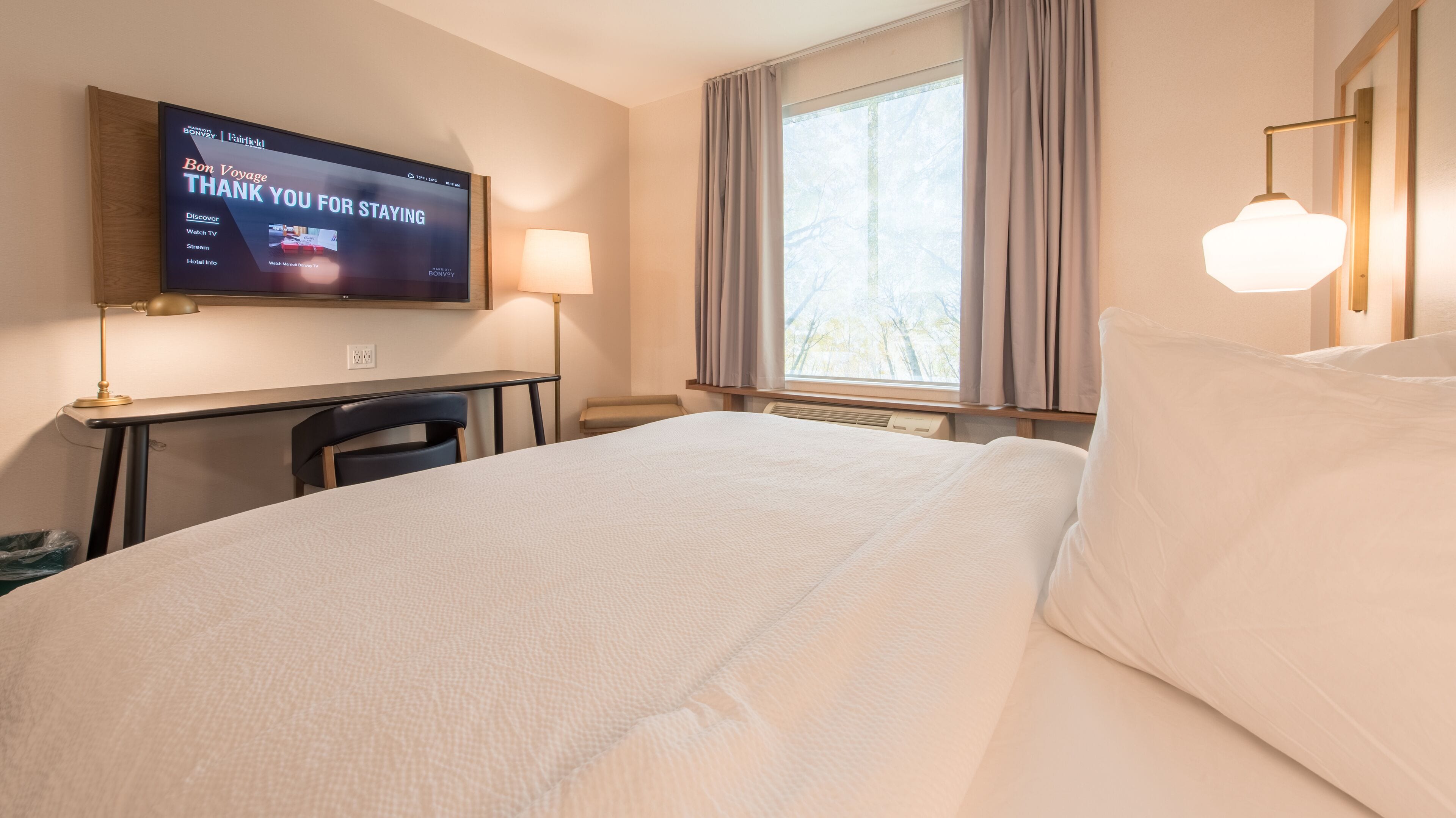 A hotel room with a bed, a desk, a TV displaying a message, a lamp, and a window with curtains.