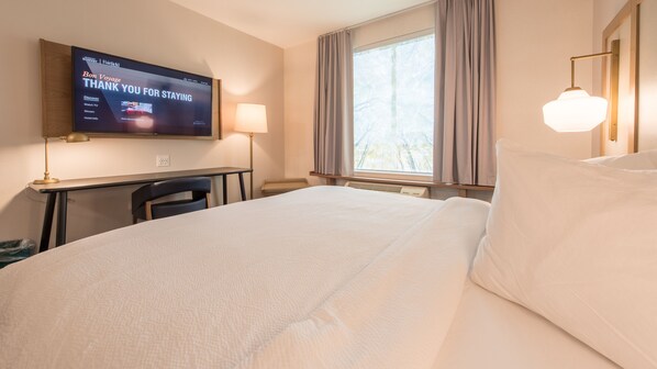 Quarto de hotel com uma cama, uma escrivaninha, uma TV exibindo uma mensagem, um abajur e uma janela com cortinas.