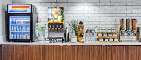A self-service beverage station with a refrigerator, a colorful fruit juice dispenser, and a coffee machine.