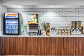 Una estación de bebidas de autoservicio con un refrigerador, un dispensador de zumos de frutas de colores y una máquina de café.