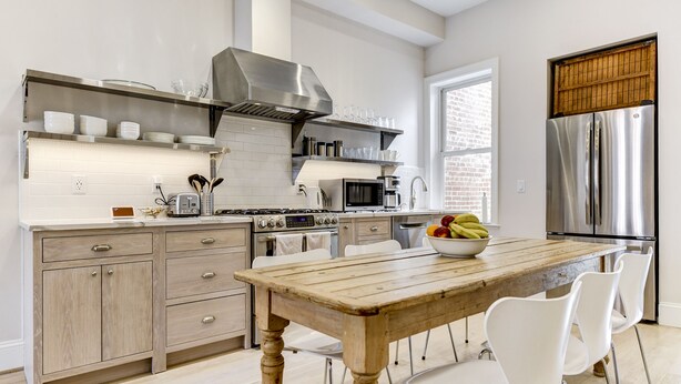 Una cocina moderna con una mesa de madera, sillas blancas, electrodomésticos de acero inoxidable y una pared de ladrillo visible a través de la ventana.