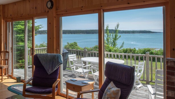 A wooden deck with two chairs and a table, overlooking a body of water and forested land.