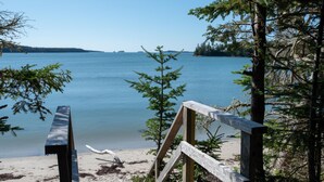 A wooden deck leading to a sandy beach with a calm body of water and trees in the background.