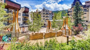 Un patio de hotel con un parque infantil, mobiliario exterior y montañas al fondo.