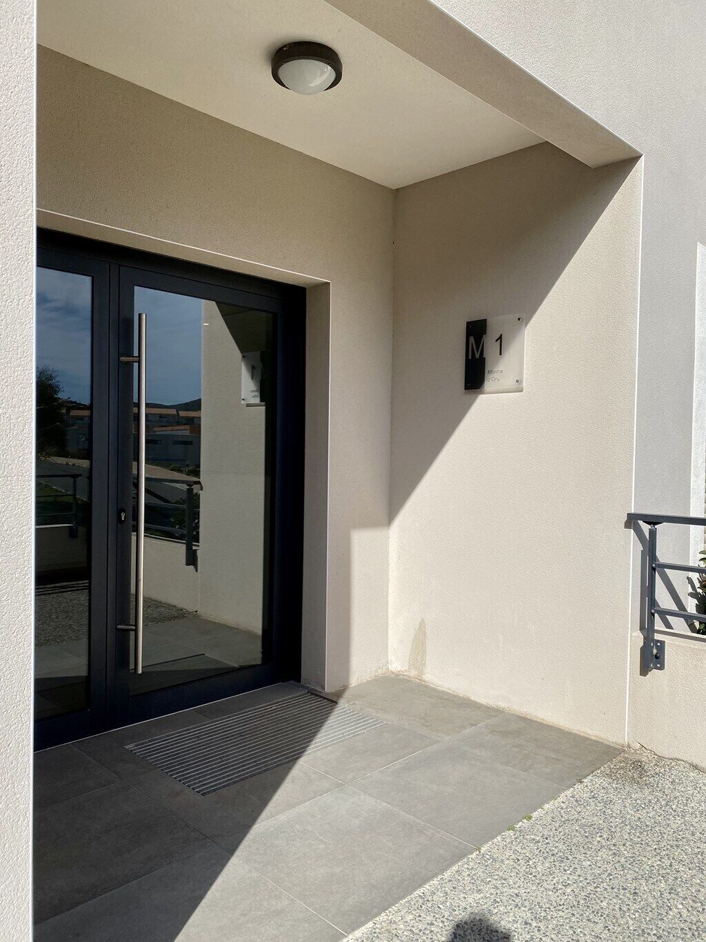 Une entrée moderne avec une porte en verre, une plaque signalétique en métal et un petit espace extérieur agrémenté d’une rambarde en métal.