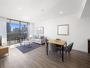 A modern living room with a sofa, dining table, and chairs. There is a large window with a city view. - Lucid Living Brisbane (South Brisbane)