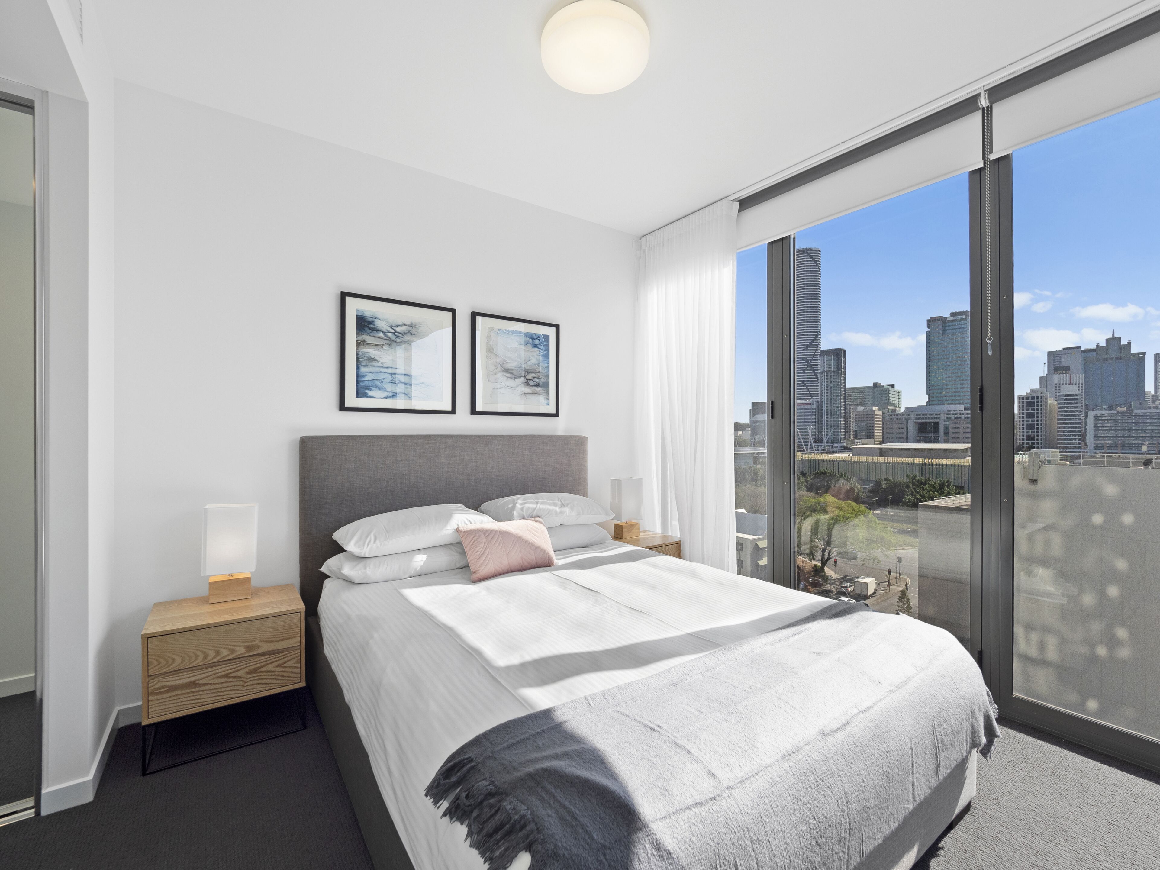 A modern bedroom with a large bed, a nightstand, a bedside table, and a city view through a large window.