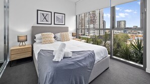 A modern bedroom with a large bed, two framed artworks, a nightstand, and a view of high-rise buildings through large windows. - Lucid Living Brisbane (South Brisbane)
