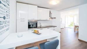 A modern kitchen with white cabinetry, a built-in oven, and a dining area with a white table and blue chairs. - Apartment Blue Chill - One-Bedroom Apartment (Mlini)