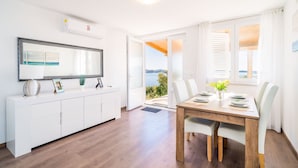 A modern dining area with a wooden table and white chairs, a white console with a mirror and decorative items, and a view of the outdoors through a glass door. - Apartment Blue Chill - One-Bedroom Apartment (Mlini)