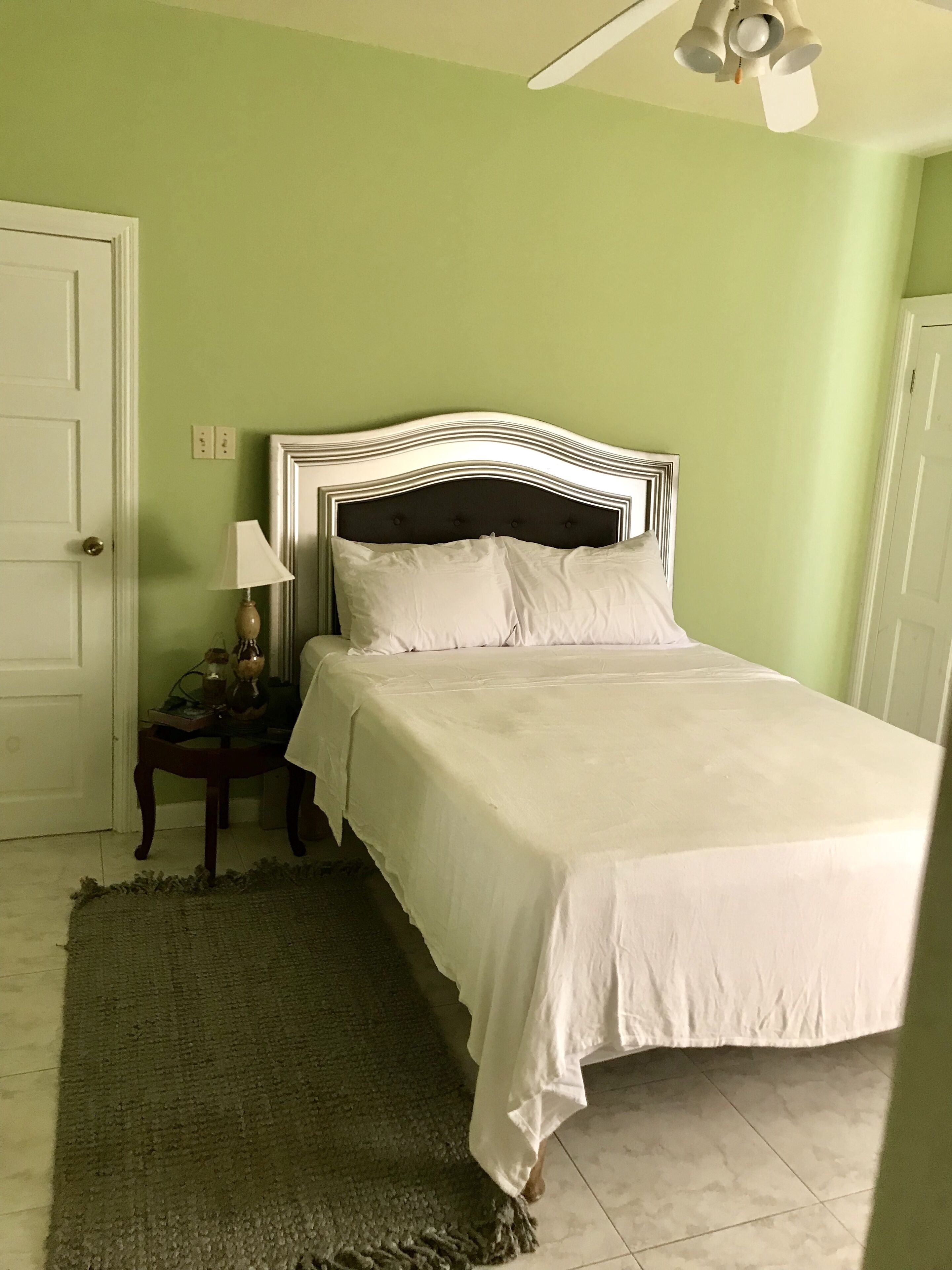 Ein ordentlich bezogenes Bett in einem Zimmer mit grüner Wand, einem weißen Deckenventilator und einem kleinen Tisch mit Lampe.
