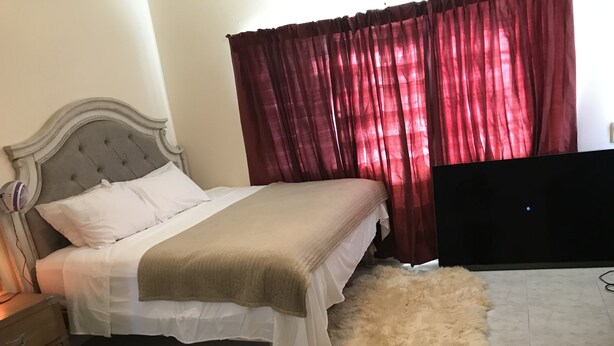 Een slaapkamer met een bed, een zacht vloerkleed, een houten nachtkastje, een televisie en rode gordijnen.
