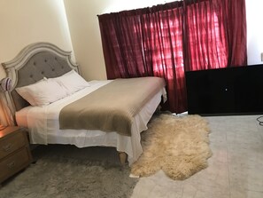 A bedroom with a bed, a fluffy rug, a wooden nightstand, a TV, and red curtains.
