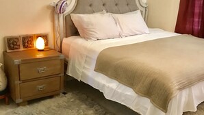 Een net opgemaakt bed met witte lakens en een bruine plaid, een zacht tapijt en een nachtkastje met een lamp en decoratieve objecten.